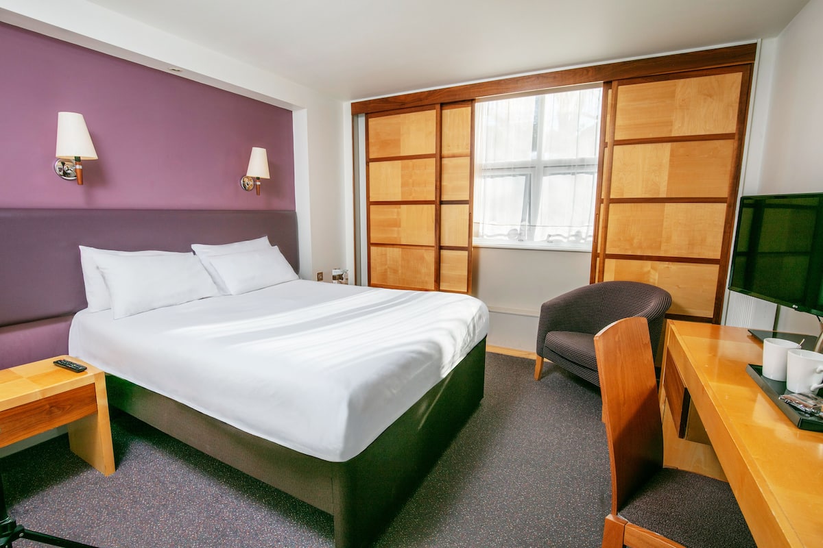 A hotel room with a bed, a desk with a chair, a window, and a TV.