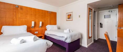 A hotel room with two beds, a wooden headboard, a purple accent wall, and a door leading to a bathroom.
