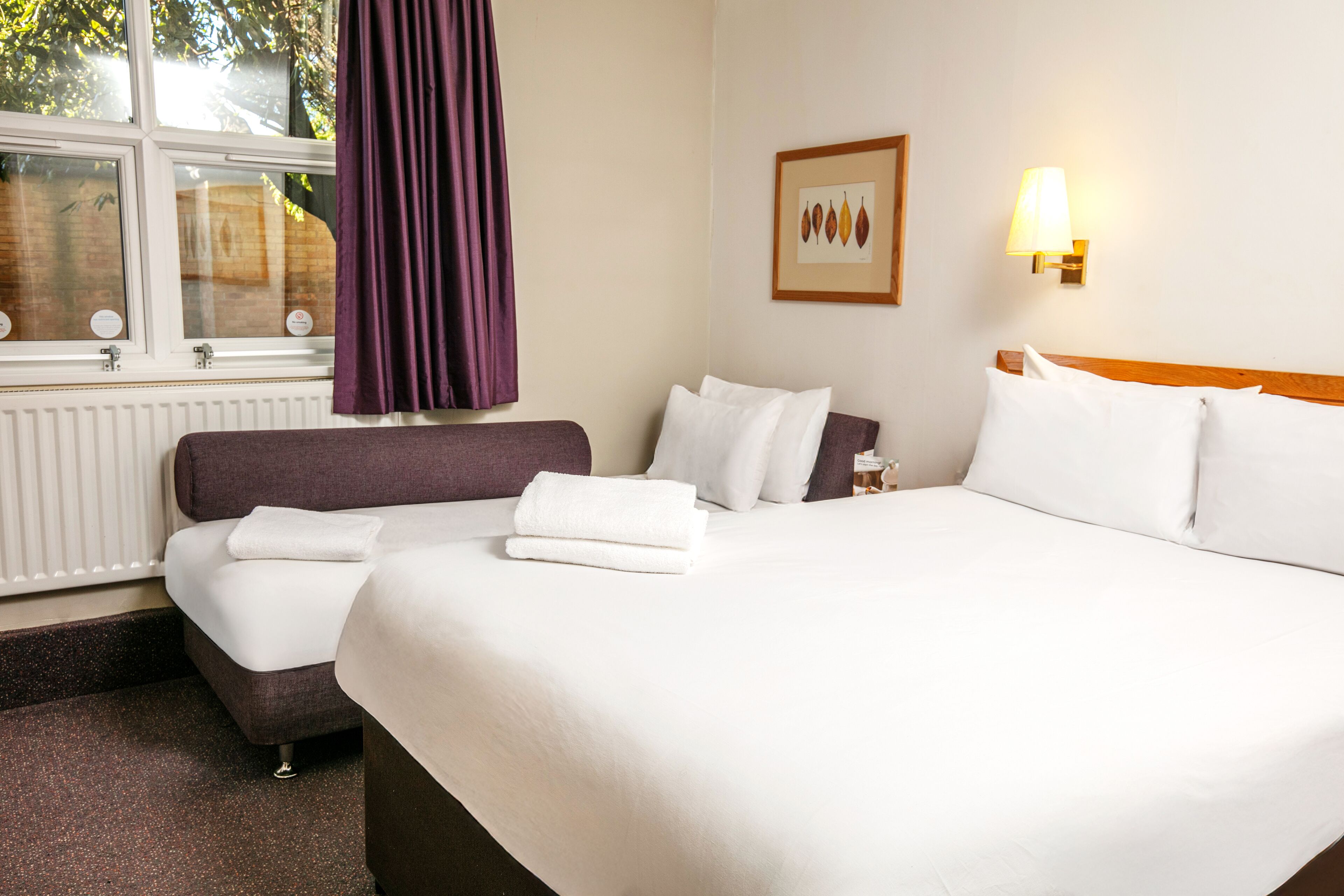 A hotel room with a bed, a sofa, a window with curtains, a wall-mounted lamp, and a framed picture.