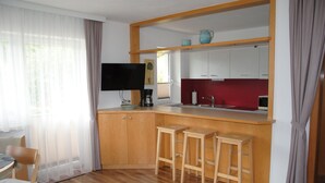 A modern kitchen with wooden cabinets, a built-in bar area with stools, a flat-screen TV, and a window with sheer curtains. - holiday apartment Elferblick (Fulpmes)