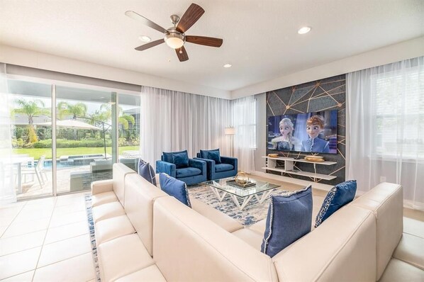 A modern living room with a beige sofa, blue armchairs, a glass coffee table, and a ceiling fan.