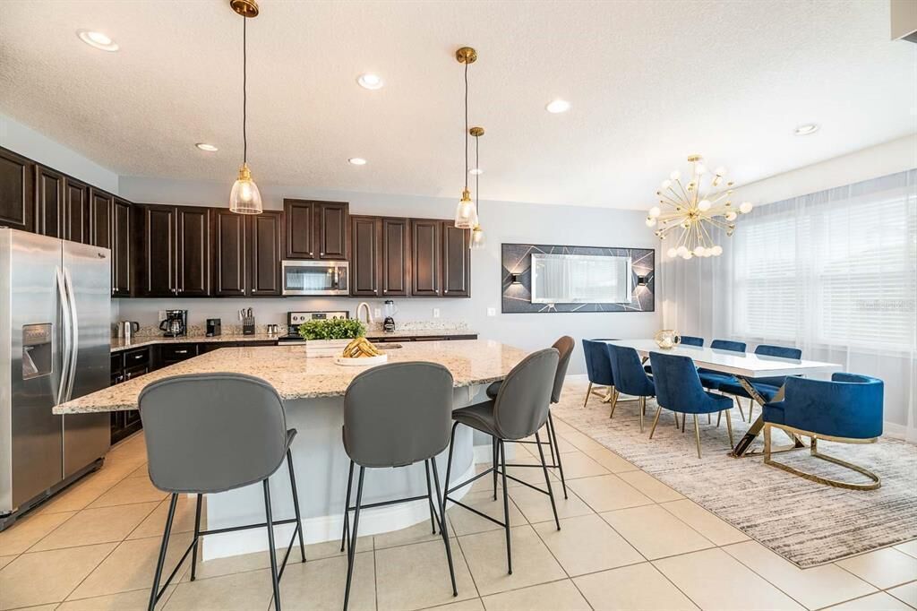 A modern kitchen with dark cabinets, a central island with bar stools, and a dining area with blue chairs and a white table.