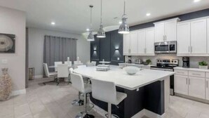 Una cocina moderna con armarios blancos, una isla central, iluminación colgante y un comedor con sillas blancas.