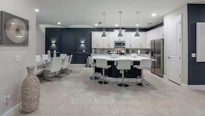 Una cocina moderna con armarios blancos, una isla central con taburetes altos, iluminación colgante y un jarrón decorativo grande.