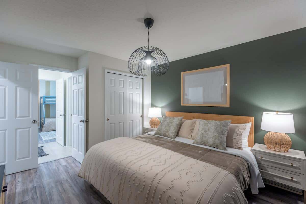A bedroom with a bed, bedside tables, a hanging light fixture, and a door leading to another room.