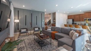 Ein modernes Wohnzimmer mit einem grauen Sofa, einem Couchtisch und einem gemusterten Teppich. Eine Treppe führt auf eine obere Ebene.