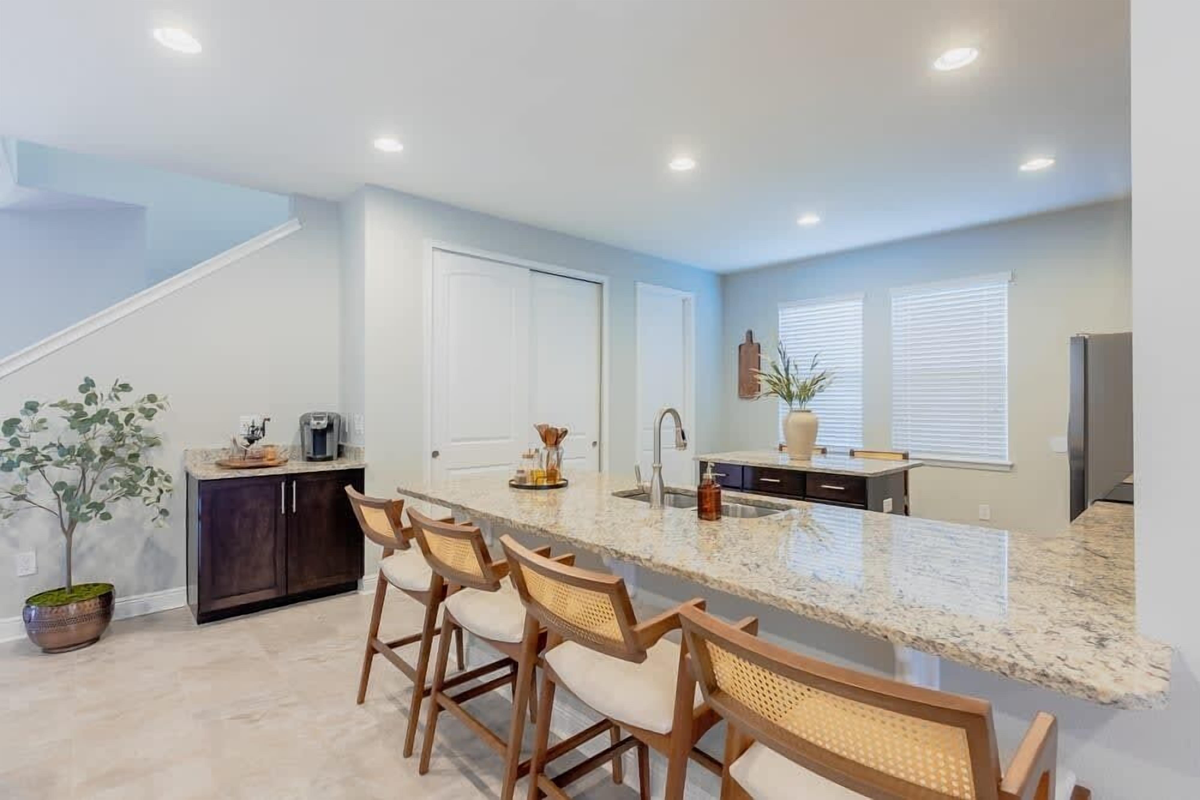 A modern kitchen with a central island, bar stools, dark cabinets, and a potted plant.