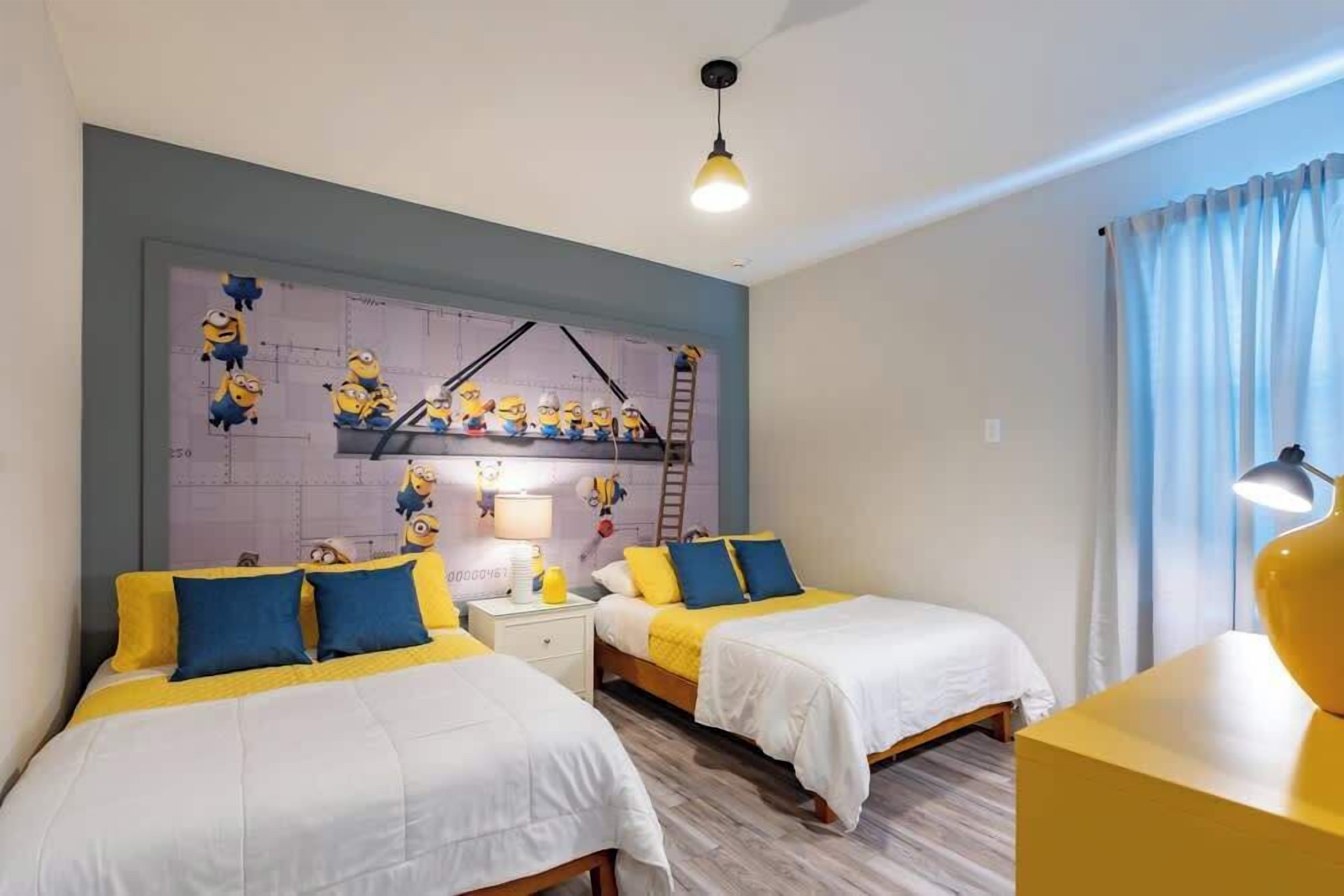 Een hotelkamer met twee bedden, een muurschildering van Minions, een ladder en een bureau met een lamp.