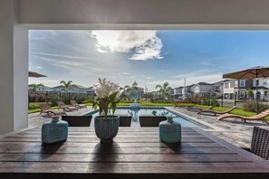 Zona junto a la piscina con mesa de madera, dos plantas en macetas y sillas de descanso.