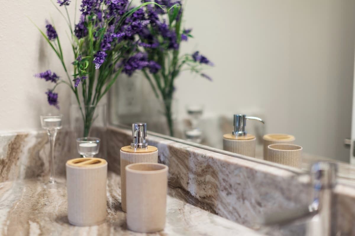 Une salle de bain avec un lavabo en marbre, des récipients cylindriques en bois et un vase rempli de fleurs violettes.