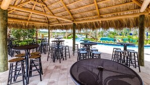 Un espace extérieur aménagé en terrasse, doté d’un toit de chaume, de plusieurs tables et de tabourets, et offrant une vue sur une piscine.