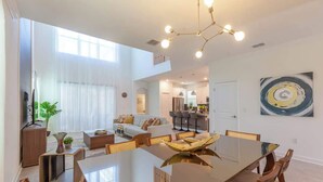 Un vaste espace dédié à la salle à manger, avec une longue table et des chaises, un lustre moderne, et une vue sur le salon et la cuisine en arrière-plan.