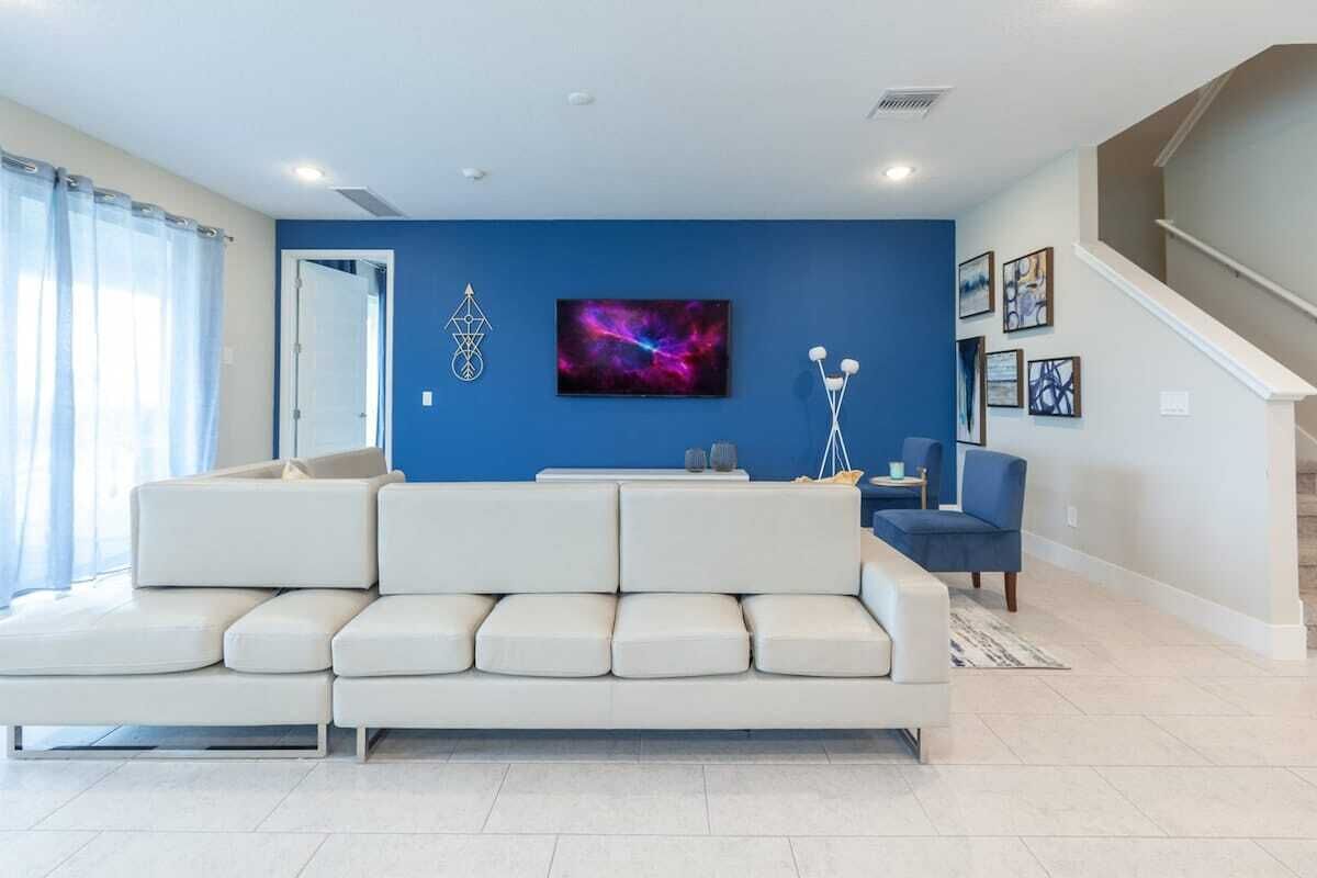 Een woonkamer met een witte modulaire bank, een blauwe accentmuur, een flatscreen televisie en een trap aan de rechterkant.