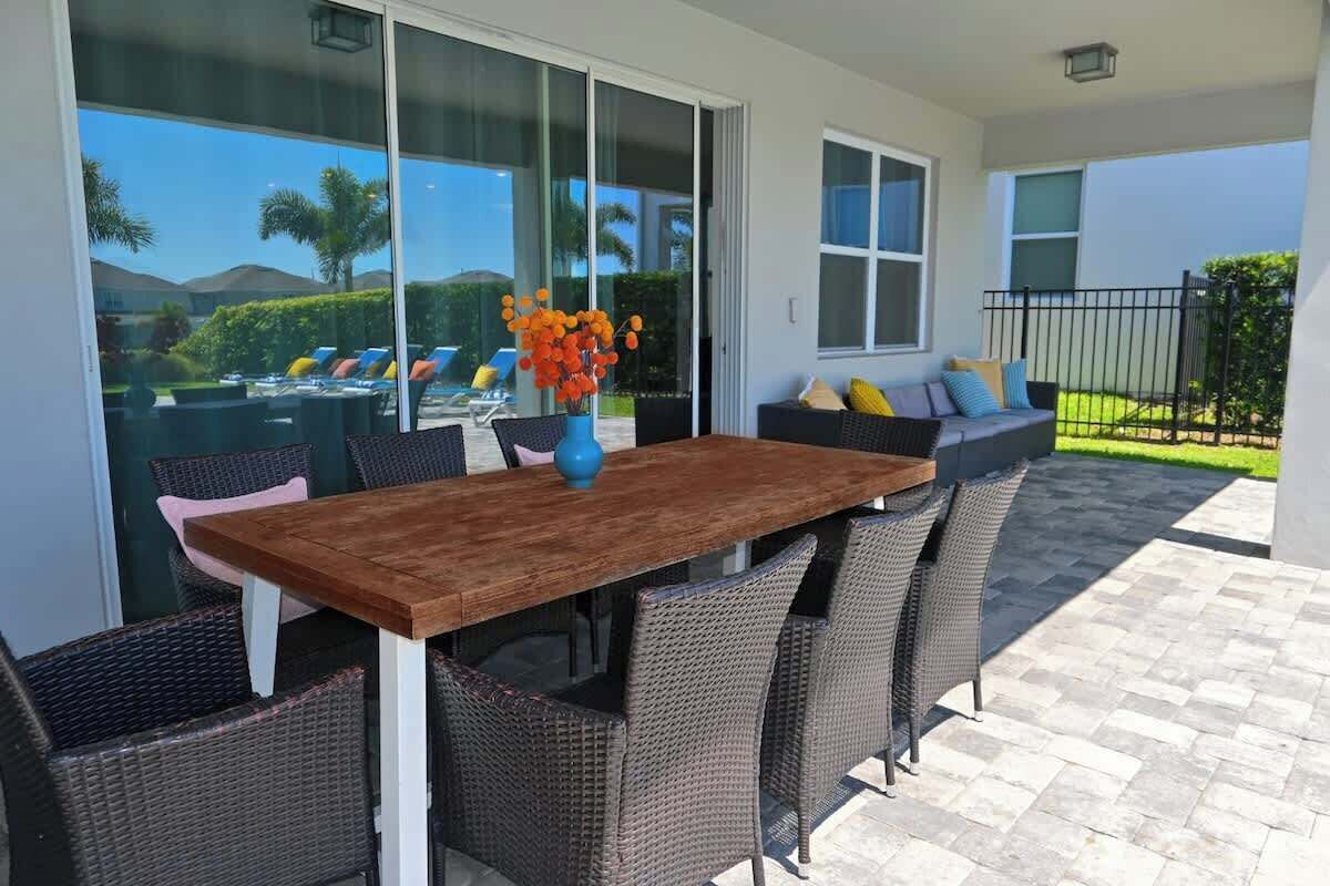 Un coin repas avec une table en bois et des chaises en osier, de grandes fenêtres et une vue sur une piscine et la verdure.