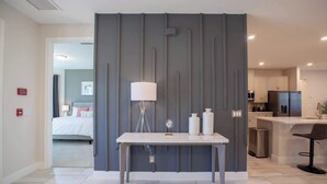Une chambre d’hôtel moderne avec un mur d’accent gris, un bureau blanc avec une lampe et une vue sur la cuisine.