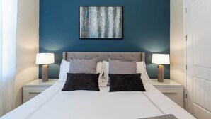 Une chambre d’hôtel avec un grand lit, deux tables de chevet avec des lampes, un mur d’accent bleu et une œuvre d’art encadrée au-dessus du lit.