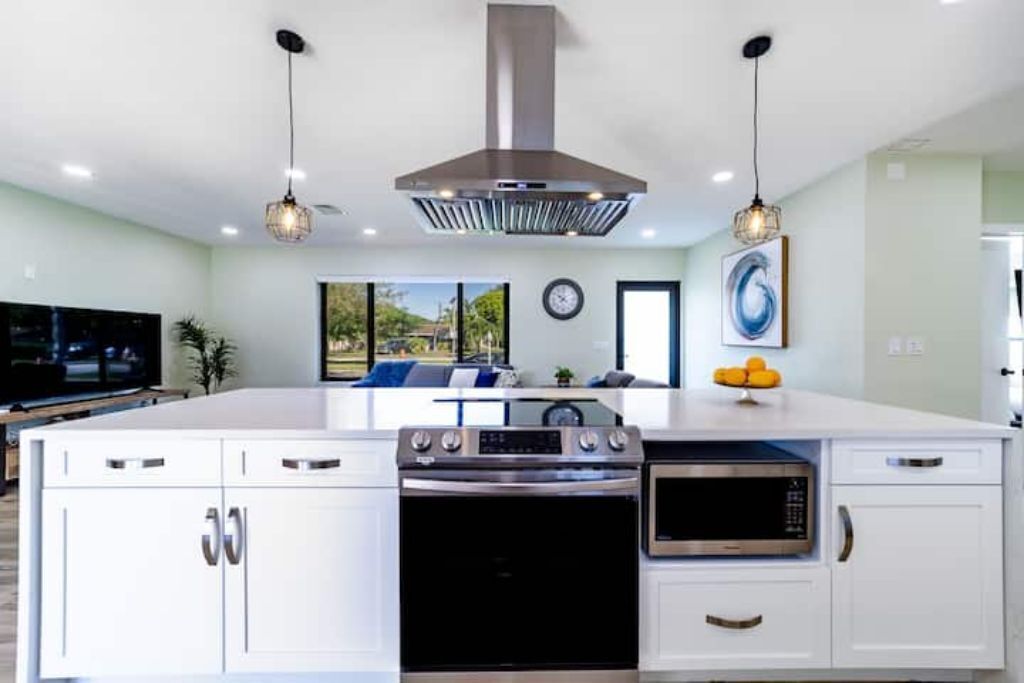 A modern kitchen with white cabinetry, a central island with appliances, pendant lighting, and a view to the living area.