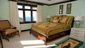 A bedroom with a bed, a chair, a nightstand, a window with curtains, and a view of the outdoors. - Maison de La Mer (Castries)