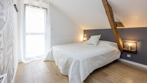 Un dormitorio con techo inclinado, una cama con ropa de cama blanca, dos lámparas de noche y una ventana con cortinas de gasa.