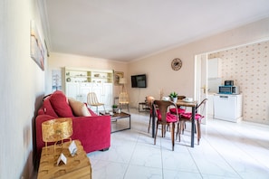 A living room with a red sofa, wooden coffee table, wicker chairs, and a dining table with chairs. A television is mounted on the wall, and a kitchen area is visible in the background. - La Brise Marine - Appartement avec balcon à 50m de la plage (Arcachon)