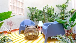 Un rincón al aire libre acogedor con dos sillas azules, una mesa de mimbre y varias macetas con plantas.