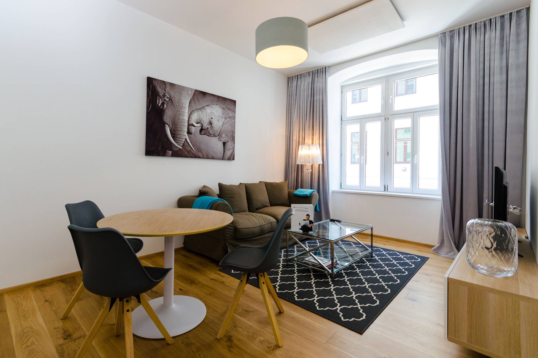 A modern living room with a round dining table, black chairs, a brown sofa, a glass coffee table, a lamp, and a large window with curtains.