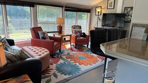 Un salon comprenant un canapé, des fauteuils, une table basse et un téléviseur. De grandes fenêtres sont équipées de stores, et on y trouve également un tapis et une lampe.