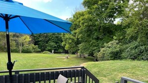 Une terrasse dotée d’une table et de chaises à plateau en verre, entourée d’une balustrade en bois, donnant sur une cour verdoyante avec des arbres.
