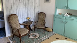 Ein Zimmer mit zwei Holzstühlen, einem kleinen Tisch mit Blumenvase, einem gemusterten Teppich und einem türkisfarbenen Schrank mit Schubladen.