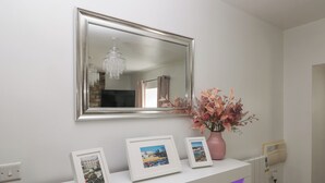A white console table with three framed photographs, a pink vase with flowers, and a mirror above. - The Old Farm House (Tenby)