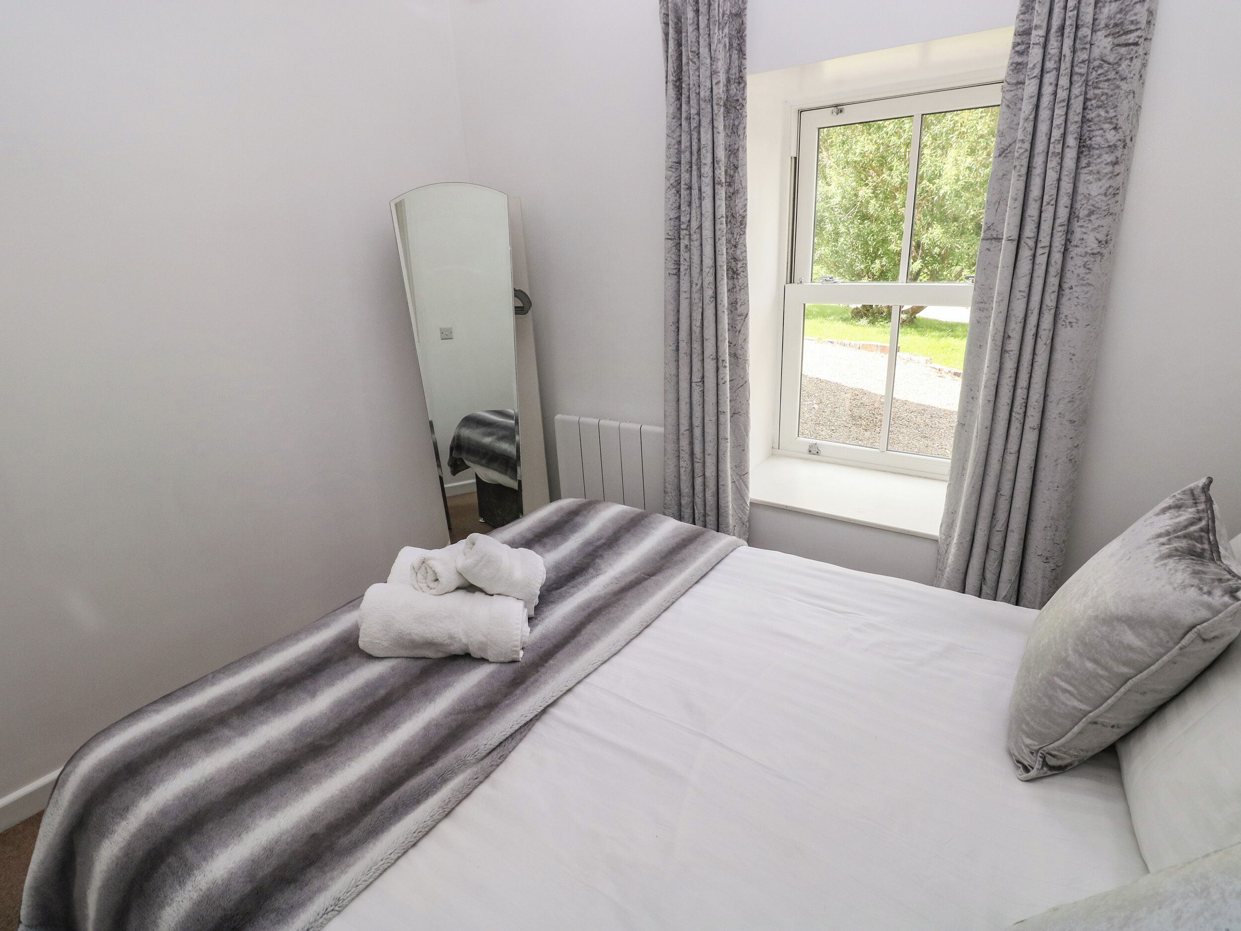 A neatly made bed with a striped blanket and two rolled towels, a window with curtains, and a mirror on the wall.