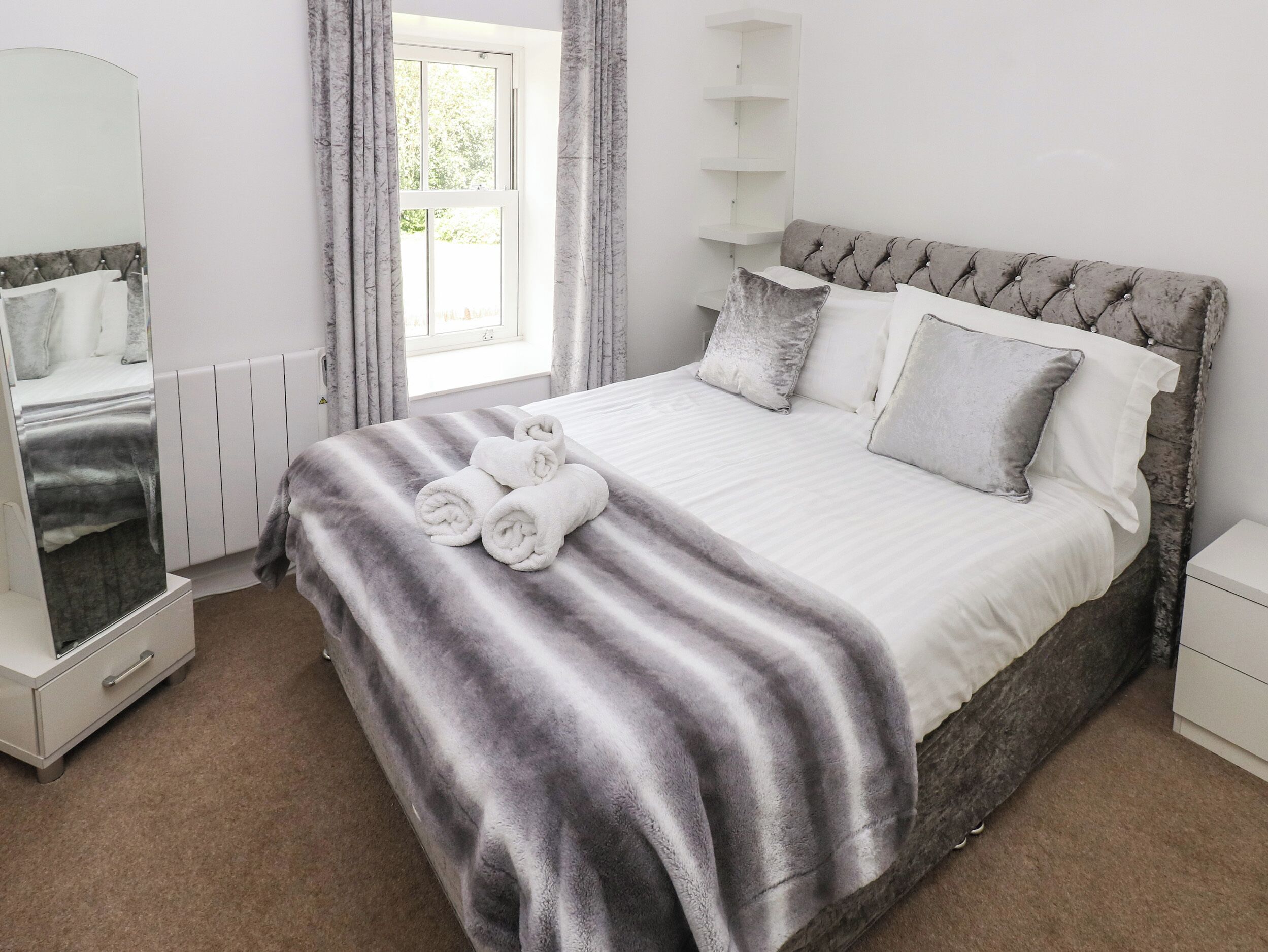 A neatly made bed with a striped blanket, a bedside table, a mirror, and a window with curtains.