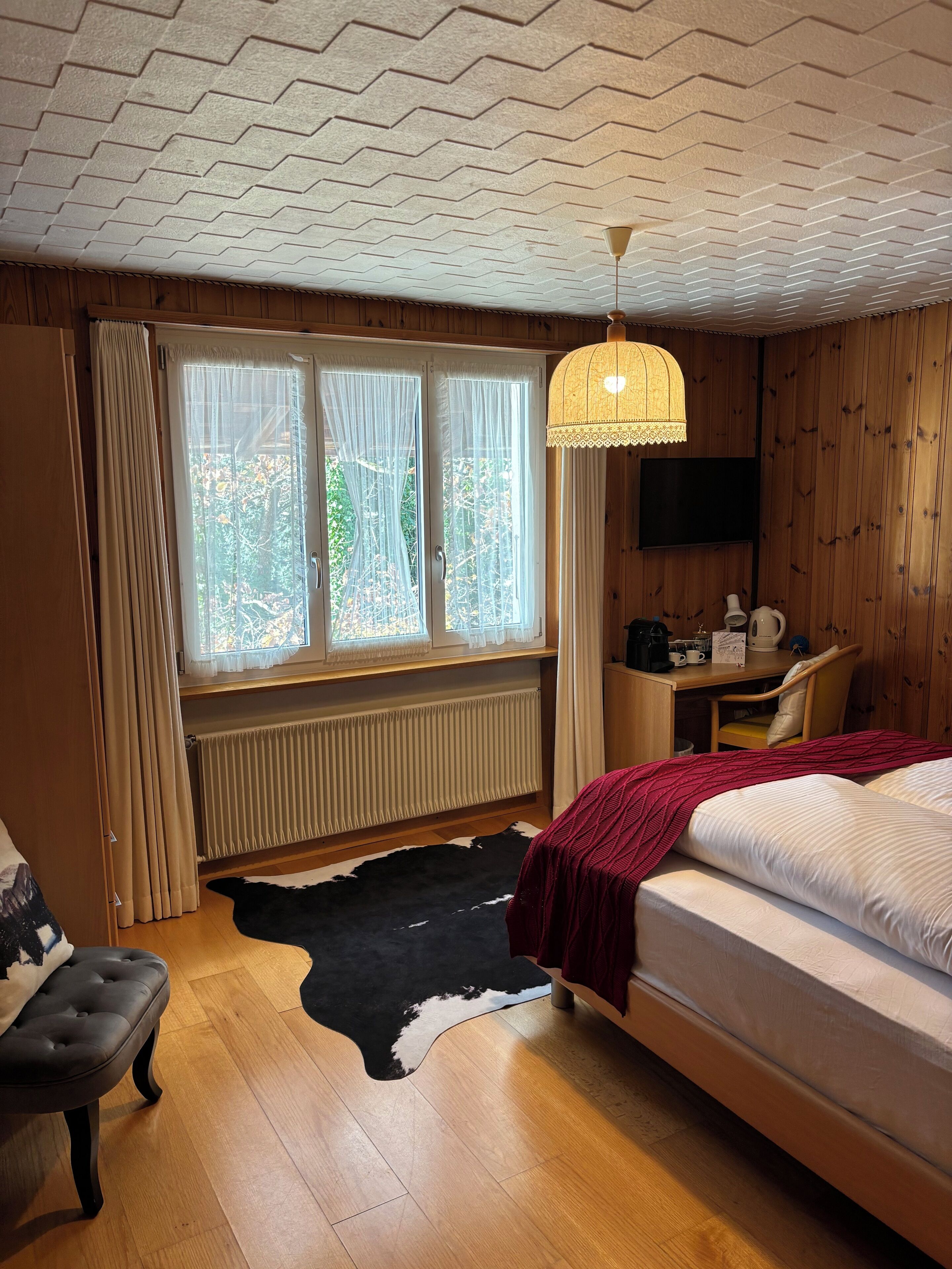 Un dormitorio con cama, escritorio, silla, radiador, alfombra de cuero de vaca y una ventana con cortinas.