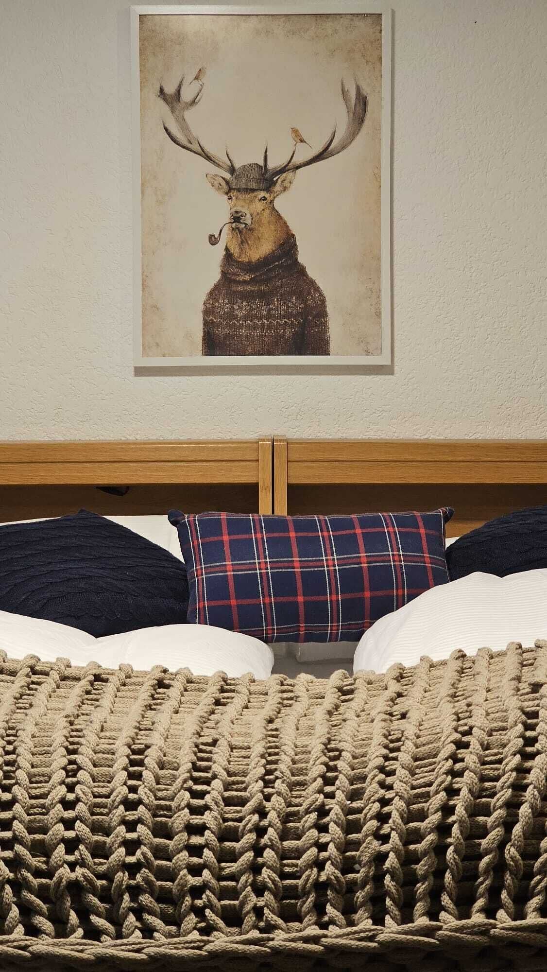 Een bed met een gebreide plaid, een ruitkussen en een lijst met een foto van een hert op de muur.