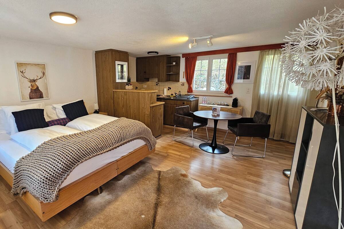 Un dormitorio con cama de madera, una mesita de noche pequeña, un tapete grande, un área de cocina con mesa y sillas, y una ventana con cortinas.