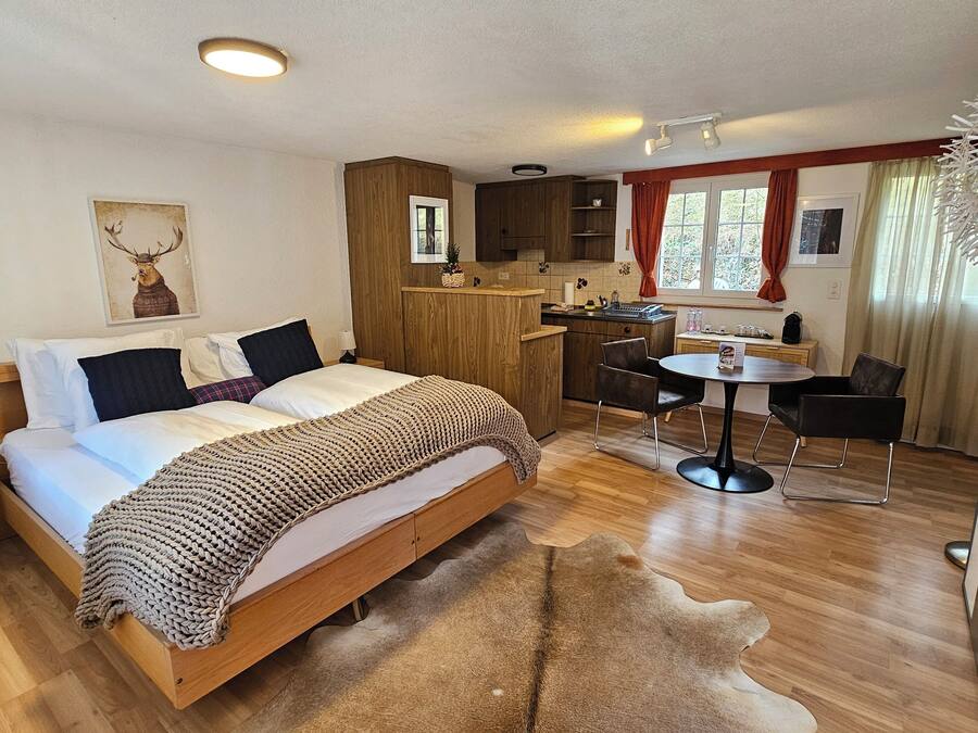 Um quarto com cama de madeira, uma pequena mesa de cabeceira, um tapete grande, um espaço de cozinha com mesa e cadeiras, e uma janela com cortinas.