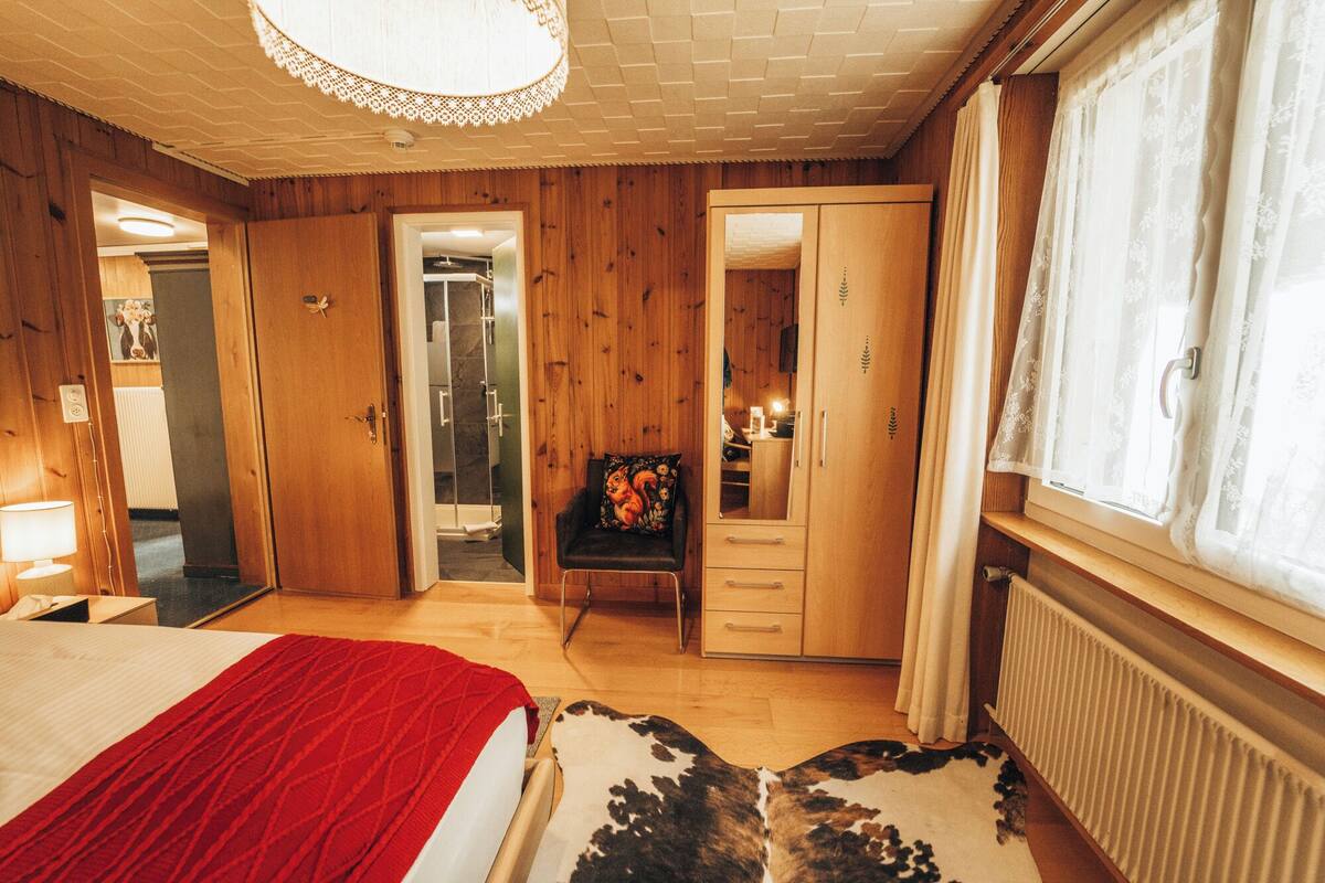 Un dormitorio con cama, espejo, armario, puerta al baño y ventana con cortinas.