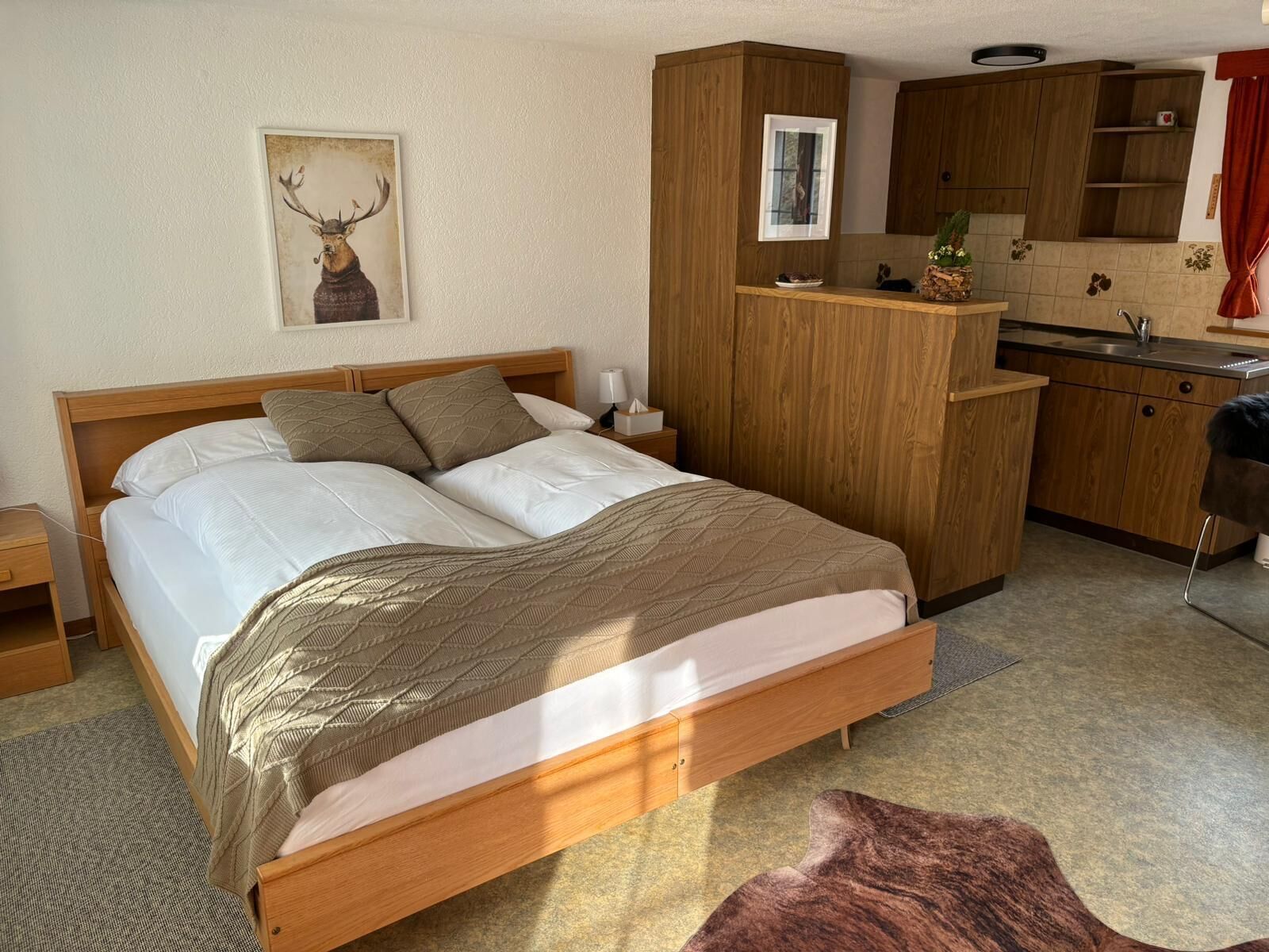 Quarto com cama de madeira, mesa de cabeceira, cozinha com armários de madeira, janela e quadro de veado fixado na parede.