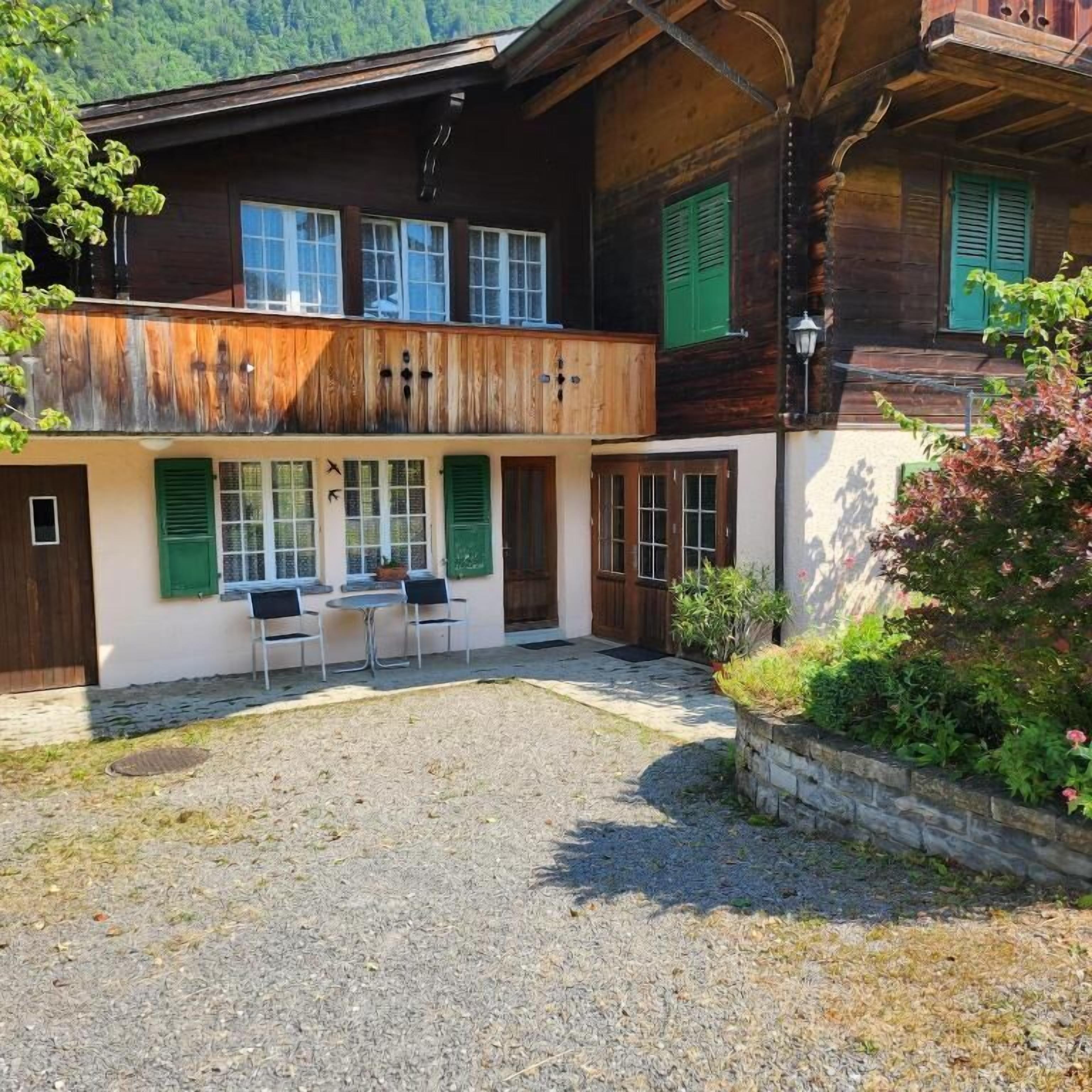 Une maison en bois à deux étages, dotée d’un balcon, d’une cour en gravier et d’un petit jardin planté.