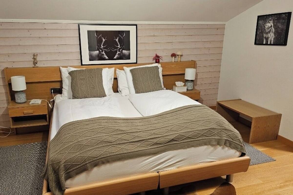 Un dormitorio ordenado con cabecera de madera, cama grande con manta estampada, dos mesitas de noche con lámparas y un cuadro enmarcado en la pared.
