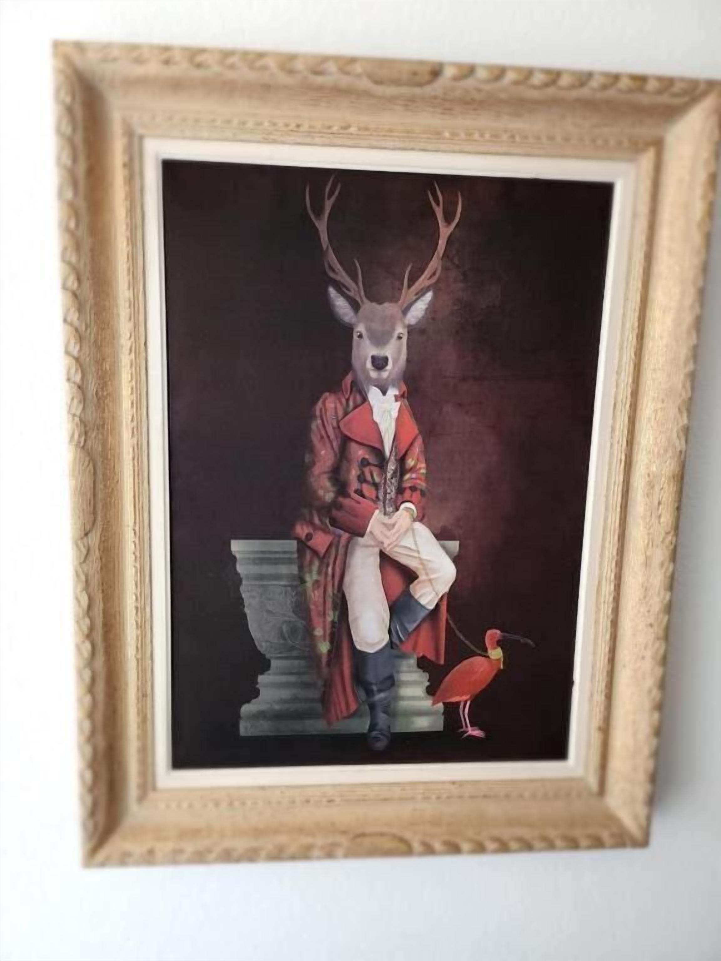Une œuvre d’art encadrée représentant un cerf portant un manteau rouge et tenant un livre, avec un oiseau perché sur son dos.