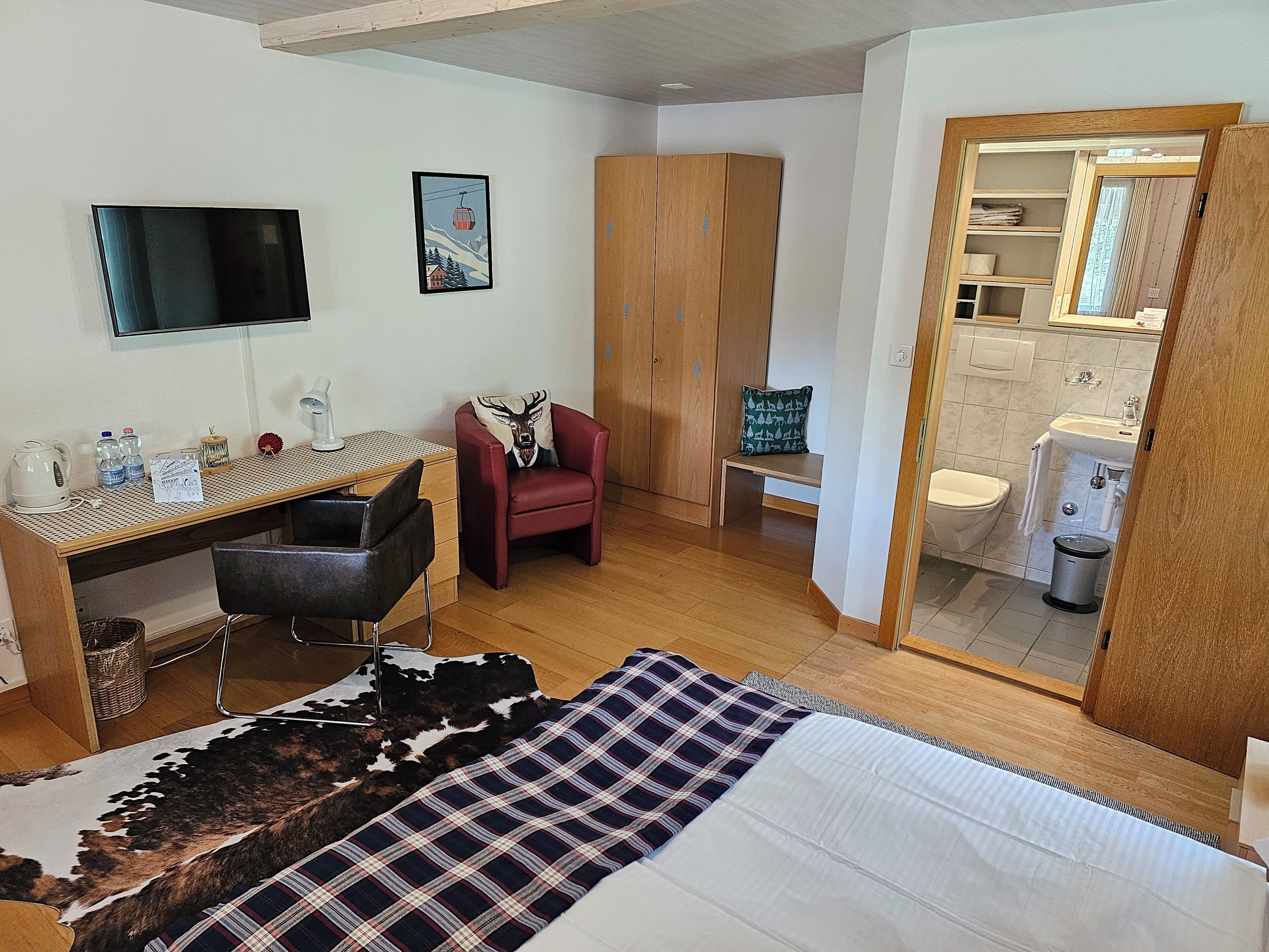 Habitación de hotel con cama, escritorio con silla, sillón rojo, pequeño banco, espejo y baño visible a través de una puerta abierta.