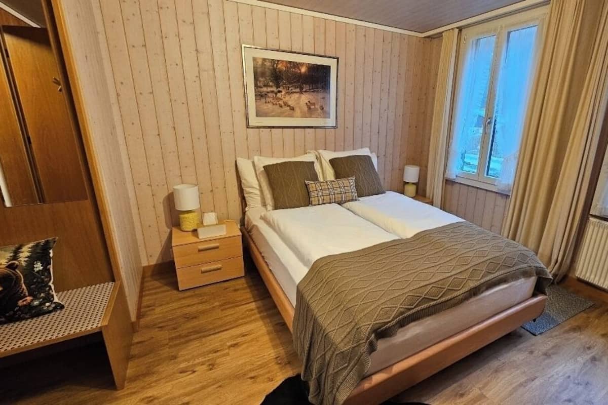 Un dormitorio con cama, mesitas de noche, una silla, un ventanal y un cuadro colgado en la pared.