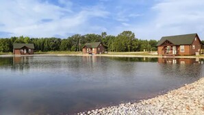 A lakeside property with three cabins, each featuring a green roof and wooden exterior, surrounded by a calm lake and a forested area. - Cabin A by the Pond - King Bed - Swimming Pool (Muscle Shoals)