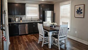 A modern kitchen with dark cabinets, stainless steel appliances, and a dining area with white chairs and a wooden table. - Cabin A by the Pond - King Bed - Swimming Pool (Muscle Shoals)