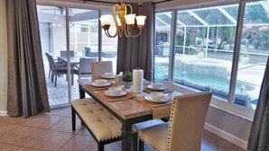 Un espace repas avec une grande table dressée pour quatre personnes, un lustre et une vue sur une piscine et un coin salon extérieur.