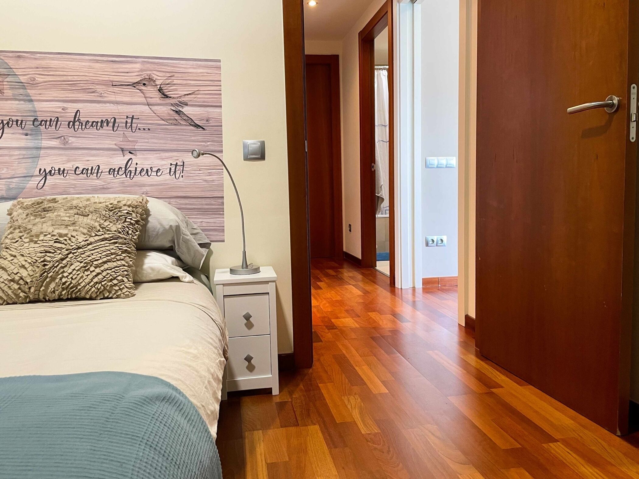 A bedroom with a bed, a bedside lamp, a wall art with motivational text, and a wooden floor.