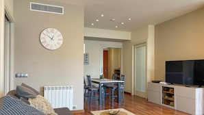 A living room with a sofa, a dining table with chairs, a TV, and a clock on the wall.