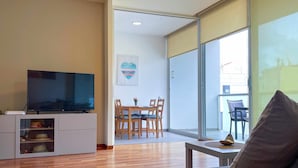 A modern living room with a flat-screen TV, wooden flooring, a grey sofa, and a dining area with wooden chairs and a table.