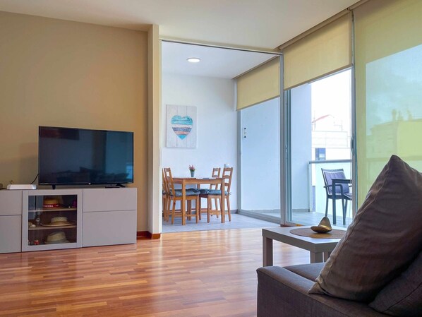 A modern living room with a flat-screen TV, wooden flooring, a grey sofa, and a dining area with wooden chairs and a table. - Walkers Paradise, Parking, Sunny Balcony - Hostem (Vilanova i la Geltrú)